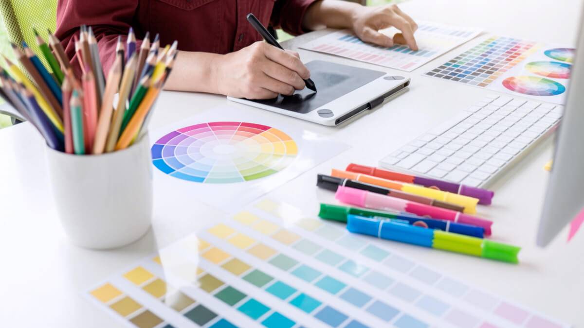 pessoa desenhando em um tablet, com a mesa repleta de canetinhas e paletas de cores