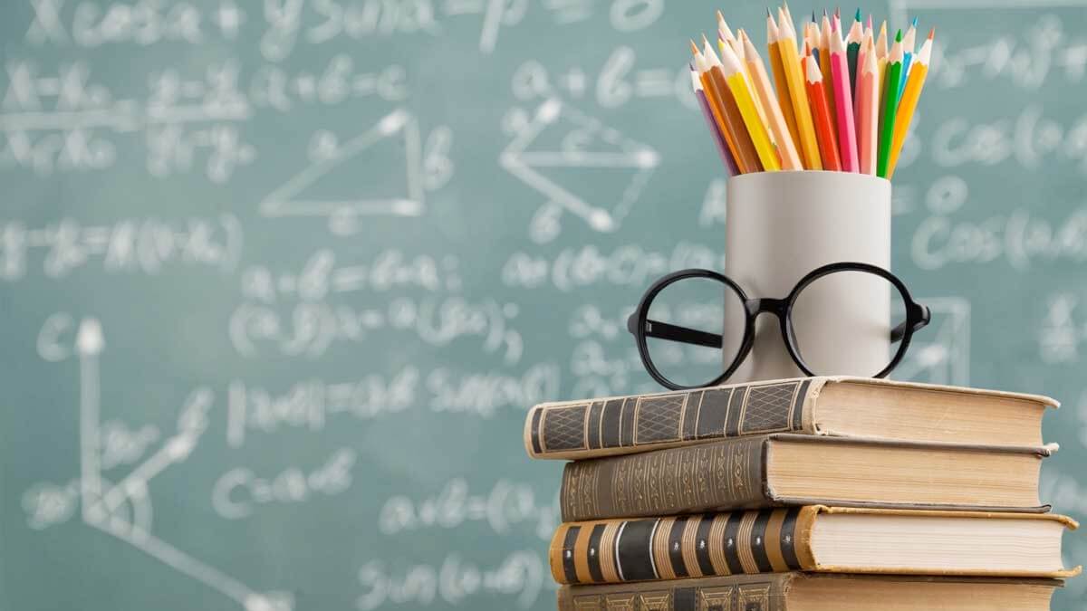 lousa de uma sala de aula; à frente, livros, um par de óculos e um pote com lápis de cor